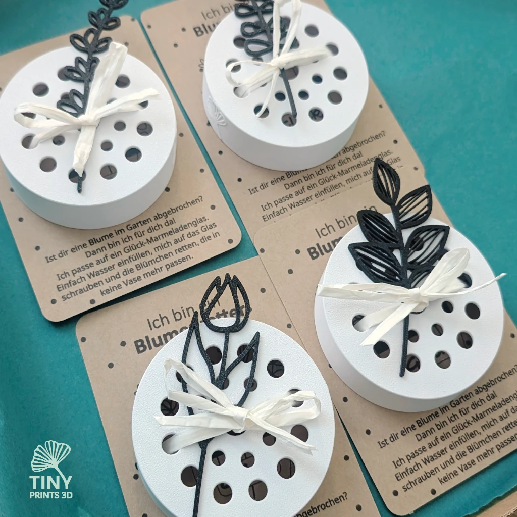 Blumenretter für das Glücksglas Kleine Stiche Vier weiße 3D-gedruckte Blumenretter mit schwarzen Blumensteckern, liebevoll auf Kraftpapier verpackt und in türkisfarbenem Seidenpapier arrangiert.