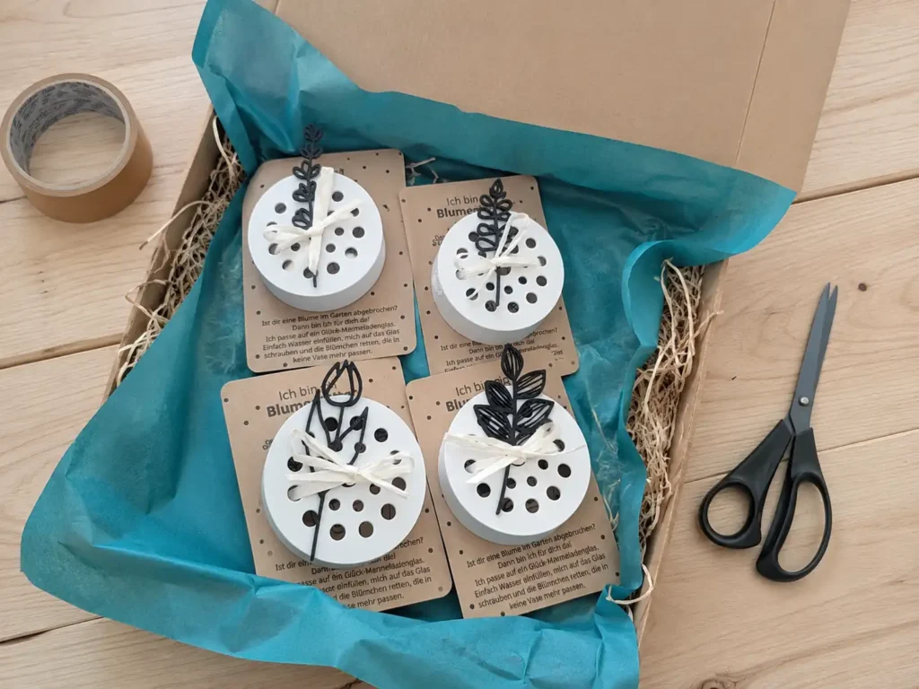 Nachhaltig verpackte Blumenretter für Marmeladengläser in einem Versandkarton mit blauem Seidenpapier, Holzwolle und Papierklebeband auf hellem Holztisch.